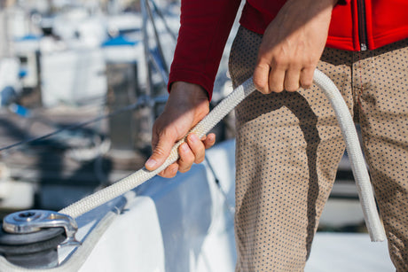 Nahaufnahme eines Menschen in roter Jacke, der an Deck eines Segelboots ein weißes Tau mit beiden Händen festhält – im Hintergrund unscharf weitere Boote im Hafen.