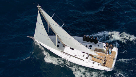 Luftaufnahme einer weißen Segelyacht mit aufgeblähten Segeln, die sich durch tiefblaues Wasser bewegt. Mehrere Personen sitzen oder stehen an Bord, während die Yacht leichte Wellen erzeugt.
