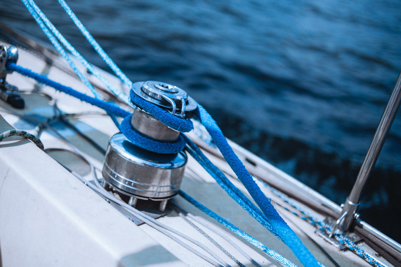 Nahaufnahme einer Winsch mit aufgewickeltem, leuchtend blauem Seil auf dem Deck eines Segelboots – im Hintergrund das tiefblaue Wasser des Meeres.