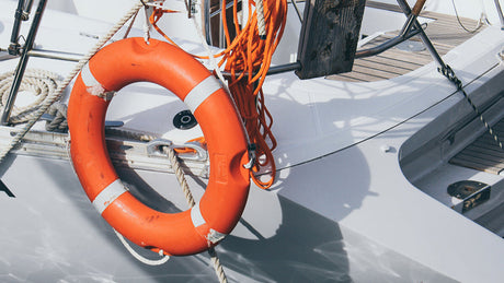 Ein orangefarbener Rettungsring mit silbernen Reflektorstreifen hängt an einem weißen Boot. Daneben ist ein Bündel orangefarbener Wurfleine befestigt, umgeben von Seilen und Reling aus Metall.