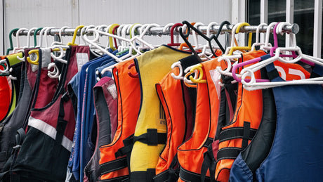 Eine Vielzahl bunter Schwimmwesten hängt dicht an dicht auf Kleiderbügeln an einer Metallstange vor einem Gebäude. Die Westen sind in verschiedenen Farben wie Orange, Blau, Gelb und Schwarz.