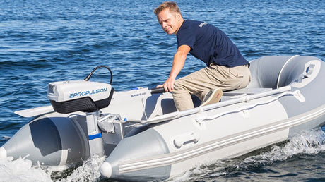 Ein Mann fährt auf einem weißen Schlauchboot mit einem Elektromotor der Marke „ePropulsion“ über ruhiges Wasser. Er trägt ein dunkelblaues T-Shirt und helle Hose und hält das Steuer des Motors in der Hand.