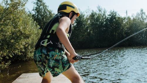 Ein Kind mit Helm, Schwimmweste und bunter Badehose steht auf einem Wakeboard am Rand eines Holzstegs und hält sich an einem Zugseil fest, bereit ins Wasser zu starten. Im Hintergrund sind Bäume und ein See zu sehen.