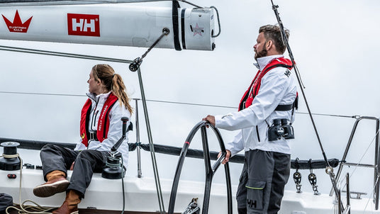 Zwei Personen auf einem Segelboot tragen weiße Segeljacken und rote automatische Rettungswesten. Eine Frau sitzt an der Seite, ein Mann steht am Steuer und hält das Steuerrad. Im Hintergrund ist ein bewölkter Himmel zu sehen.