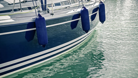 Dunkelblaues Segelboot mit mehreren länglichen, ebenfalls dunkelblauen Fendern, die entlang der Bordwand hängen und das Boot im Hafen vor Beschädigungen schützen. Das Boot schwimmt im grünen, ruhigen Wasser.