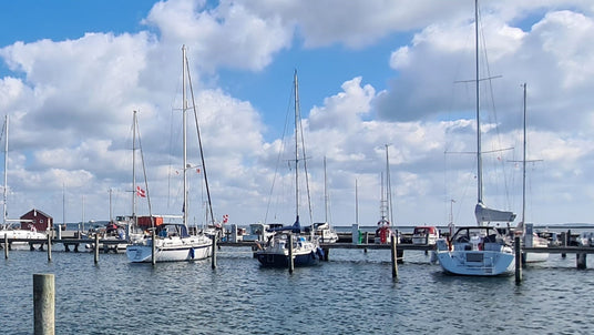 Mehrere Segelboote liegen nebeneinander in den Boxen eines Yachthafens, der von Holzstegen und Pfählen begrenzt ist. Im Hintergrund sind kleine rote Hafenhäuser und eine Zapfsäule für Bootskraftstoff zu sehen. Der Himmel ist leicht bewölkt bei sonnigem Wetter.