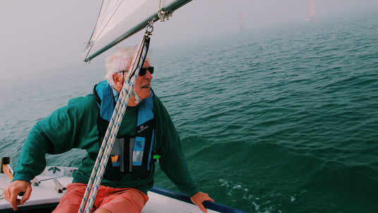 Ein älterer Mann mit weißem Haar und Sonnenbrille segelt bei Nebel auf dem offenen Meer. Er trägt eine grün-blaue Rettungsweste und sitzt aufmerksam im Cockpit eines kleinen Segelboots.