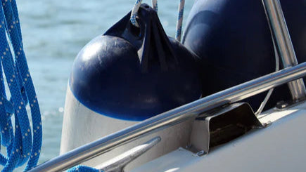 Zwei große, blaue Bootsfender hängen an einem Edelstahlgeländer eines Bootes mit Seilen daneben.