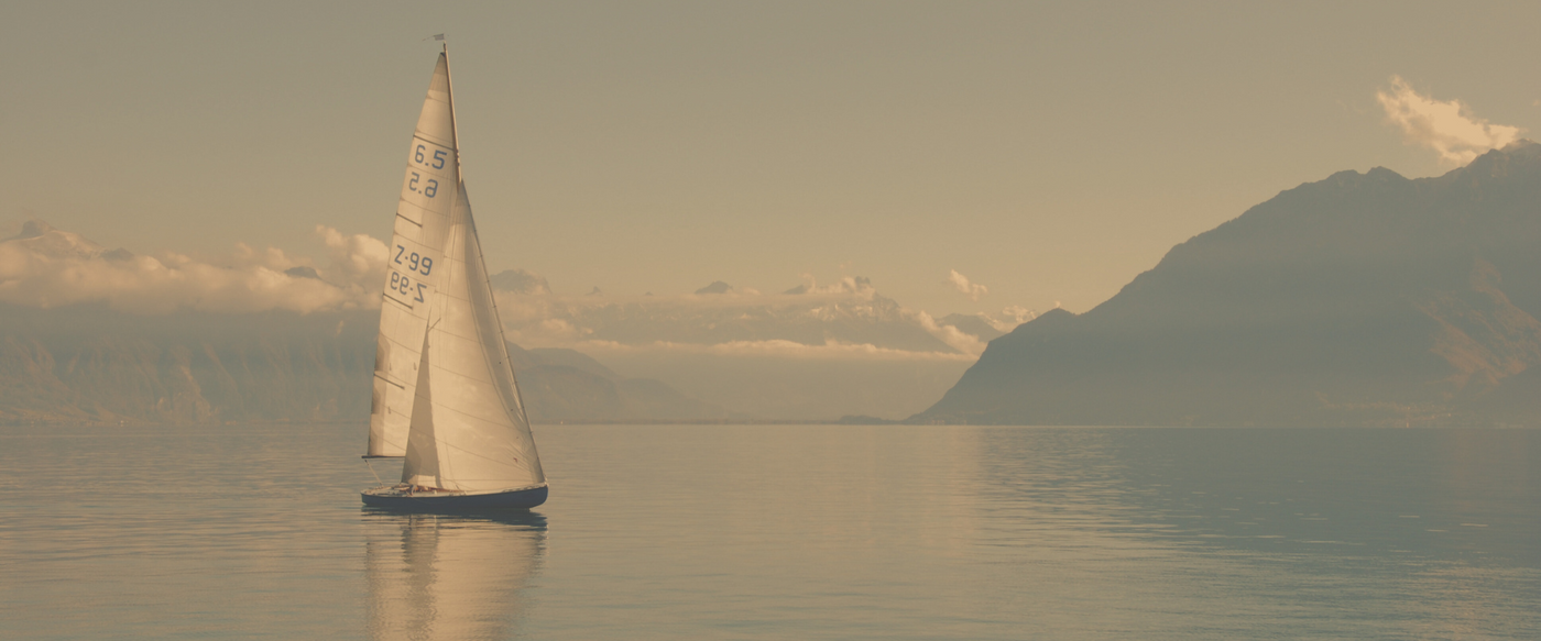Ein einzelnes Segelboot gleitet ruhig über einen spiegelglatten See, umgeben von beeindruckenden Bergen unter einem klaren, blauen Himmel. Die Szene strahlt Ruhe und Freiheit aus.