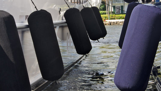 Mehrere große Yachten liegen nebeneinander im Wasser. Zwischen den Booten hängen zahlreiche zylinderförmige Bootsfender mit schwarzen und dunkelblauen Stoffüberzügen, um die Rümpfe vor Beschädigungen beim Anlegen zu schützen. Im Hintergrund sind Palmen und Gebäude zu sehen.