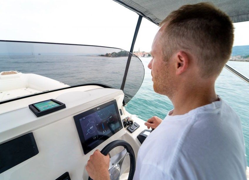 Ein Mann steuert ein Motorboot über ein digitales Display und Lenkrad. Im Hintergrund sind Meer, Küste und ein bewölkter Himmel zu sehen.