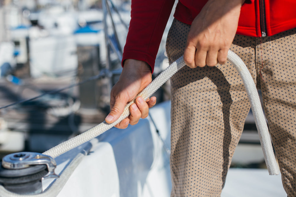 Nahaufnahme eines Menschen in roter Jacke, der an Deck eines Segelboots ein weißes Tau mit beiden Händen festhält – im Hintergrund unscharf weitere Boote im Hafen.