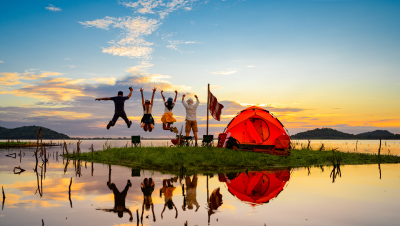 Fünf Menschen springen fröhlich in die Luft auf einer kleinen, grünen Insel im Wasser neben einem orangefarbenen Zelt. Der Sonnenuntergang spiegelt sich im ruhigen Wasser, im Hintergrund sind Berge zu sehen.