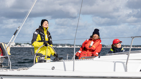 Drei Personen in wetterfester Segelkleidung auf einem weißen Segelboot bei bewölktem Himmel und ruhiger See.