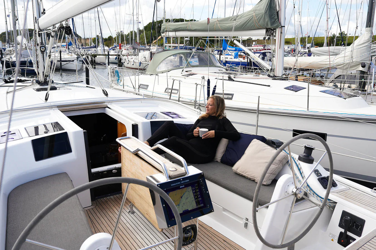 Frau sitzt entspannt mit einer Tasse Kaffee im Cockpit einer modernen Segelyacht im Hafen, umgeben von weiteren festgemachten Segelbooten.
