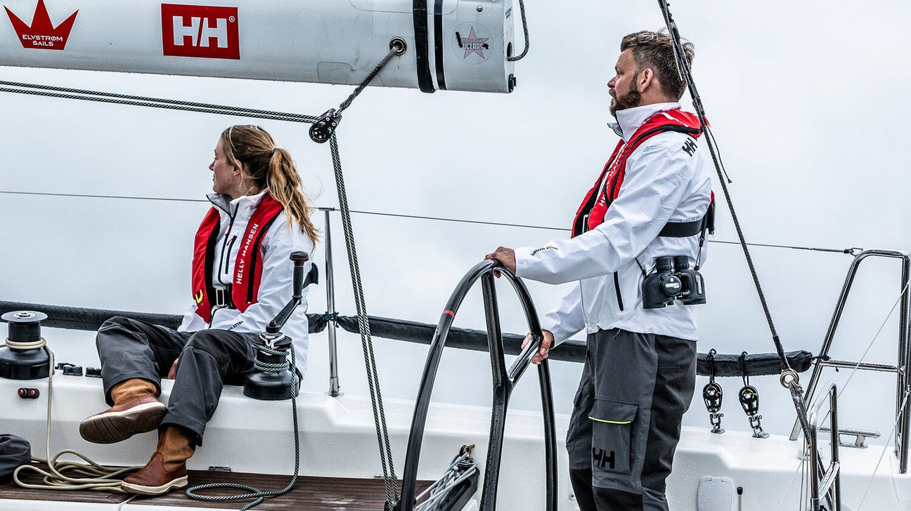 Zwei Personen auf einem Segelboot tragen weiße Segeljacken und rote automatische Rettungswesten. Eine Frau sitzt an der Seite, ein Mann steht am Steuer und hält das Steuerrad. Im Hintergrund ist ein bewölkter Himmel zu sehen.