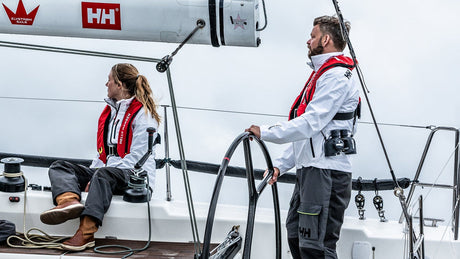 Zwei Personen auf einem Segelboot tragen weiße Segeljacken und rote automatische Rettungswesten. Eine Frau sitzt an der Seite, ein Mann steht am Steuer und hält das Steuerrad. Im Hintergrund ist ein bewölkter Himmel zu sehen.
