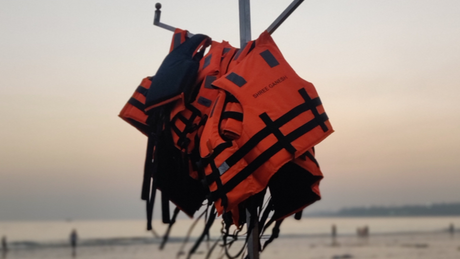 Mehrere orangefarbene Rettungswesten hängen an einem Metallständer am Strand. Im Hintergrund ist das Meer bei Sonnenuntergang zu sehen, einige Menschen stehen im flachen Wasser.