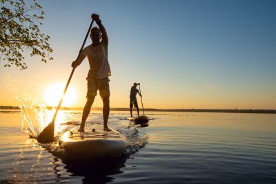 Zwei Personen fahren bei Sonnenuntergang auf Stand-Up-Paddle-Boards über ruhiges Wasser. Die vordere Person spritzt mit dem Paddel Wasser auf, beide sind im Gegenlicht als Silhouetten erkennbar.