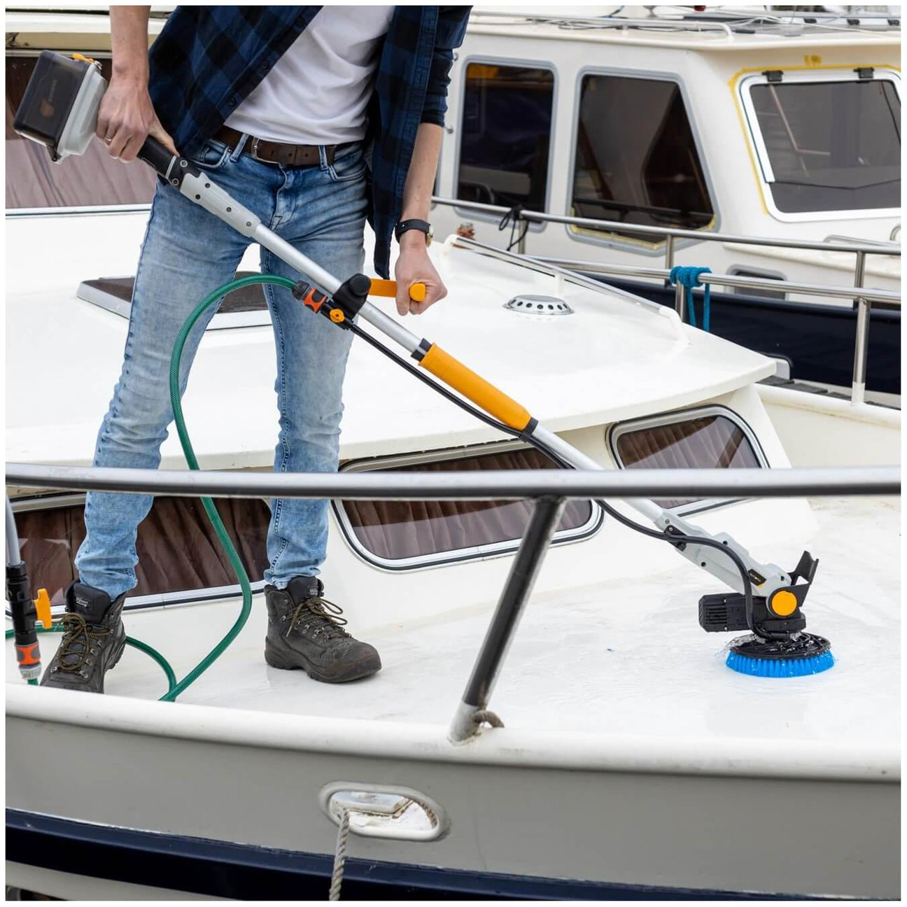 Eine Person reinigt das Deck eines Bootes mit einer elektrisch betriebenen Reinigungsbürste mit Teleskopstiel. Ein Wasserschlauch ist an das Gerät angeschlossen. Im Hintergrund sind weitere Boote im Hafen zu sehen.