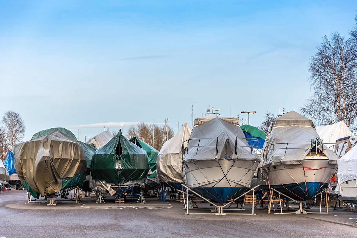 Mehrere Boote stehen im Winterlager auf einem asphaltierten Platz, mit Planen in verschiedenen Farben abgedeckt und sicher aufgebockt, um sie vor Witterung zu schützen.