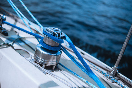 Nahaufnahme einer Winsch mit aufgewickeltem, leuchtend blauem Seil auf dem Deck eines Segelboots – im Hintergrund das tiefblaue Wasser des Meeres.
