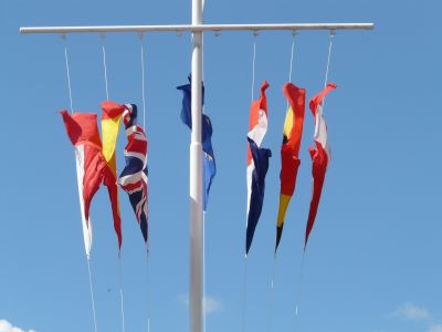 Mehrere bunte Schiffsflaggen hängen an einem senkrechten Mast mit Querträger vor blauem Himmel. Die Flaggen sind nach unten hängend und zeigen verschiedene Farben und Muster.