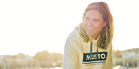 Eine lächelnde Person mit langen, hellbraunen Haaren trägt einen cremefarbenen Hoodie mit der Aufschrift „MUSTO“. Die Sonne scheint im Hintergrund, der Himmel ist hell, und unscharf sind Wasser und Gebäude zu erkennen.