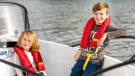 Zwei Kinder sitzen auf einem kleinen weißen Motorboot und tragen rote Rettungswesten. Sie lächeln in die Kamera, während das Boot auf ruhigem Wasser treibt. Im Hintergrund ist das Ufer mit Bäumen zu sehen.