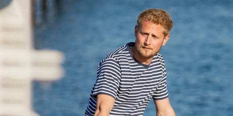 Ein Mann mit blondem, kurzem Haar und Bart trägt ein blau-weiß gestreiftes T-Shirt. Er sitzt oder steht nahe am Wasser, das im Hintergrund blau schimmert, und blickt leicht nach rechts.