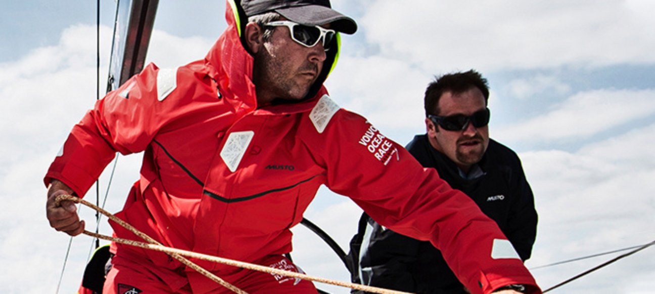 Zwei Männer auf einem Segelboot bei sportlicher Aktivität. Der vordere trägt eine rote Segeljacke mit Reflektoren, Sonnenbrille und Mütze und zieht an einem Seil. Der hintere trägt eine dunkelblaue Jacke und beobachtet die Situation konzentriert.