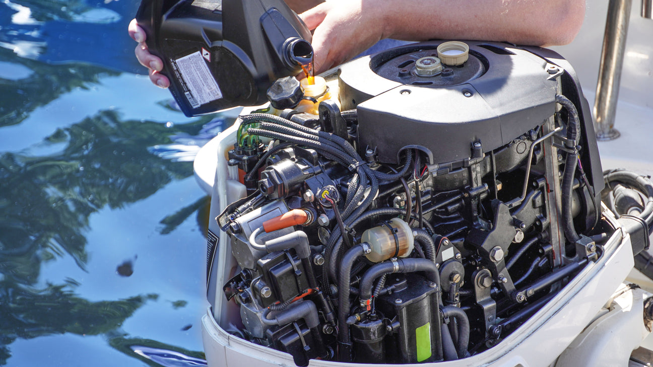 Eine Person füllt Motoröl in einen geöffneten Außenbordmotor eines Bootes. Der Motor ist detailliert sichtbar mit Kabeln, Leitungen und technischen Komponenten. Im Hintergrund spiegelt sich Wasser.