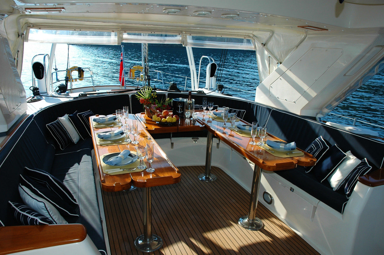 Elegant gedeckter Esstisch im geschützten Cockpit einer Yacht mit Meerblick, dekoriert mit Obstschale, Gläsern und Stoffservietten – bereit für ein stilvolles Abendessen an Bord.