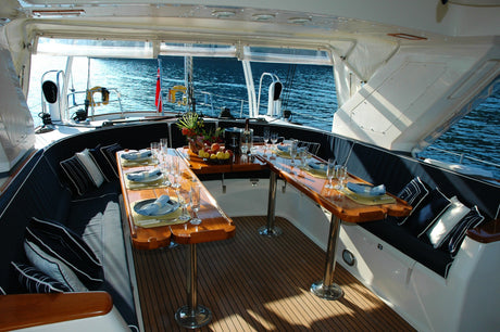 Elegant gedeckter Esstisch im geschützten Cockpit einer Yacht mit Meerblick, dekoriert mit Obstschale, Gläsern und Stoffservietten – bereit für ein stilvolles Abendessen an Bord.