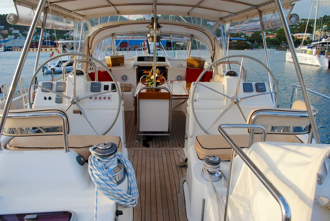 Blick in das Cockpit einer modernen Segelyacht mit zwei Steuerrädern, Navigationsinstrumenten, Sitzpolstern und einem gedeckten Tisch im Hintergrund. Das Boot liegt im Hafen, im Hintergrund sind Wasser, andere Boote und ein Ufer mit Gebäuden zu sehen.