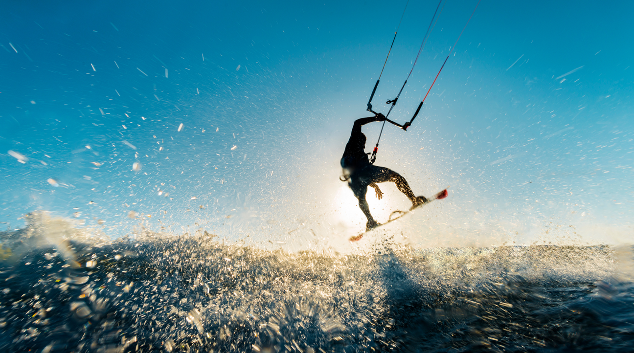 Eine Person springt dynamisch mit einem Kiteboard über das Wasser, während Sonnenlicht durch die Silhouette scheint. Wasser spritzt in alle Richtungen, der Himmel ist klar und blau.