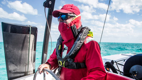 Eine Person in roter Segelkleidung und Schwimmweste steht am Steuer eines Segelboots auf offenem Meer. Sie trägt eine rote Mütze, Sonnenbrille und hat das Gesicht teilweise bedeckt. Der Himmel ist blau mit einigen Wolken, das Wasser türkisfarben.