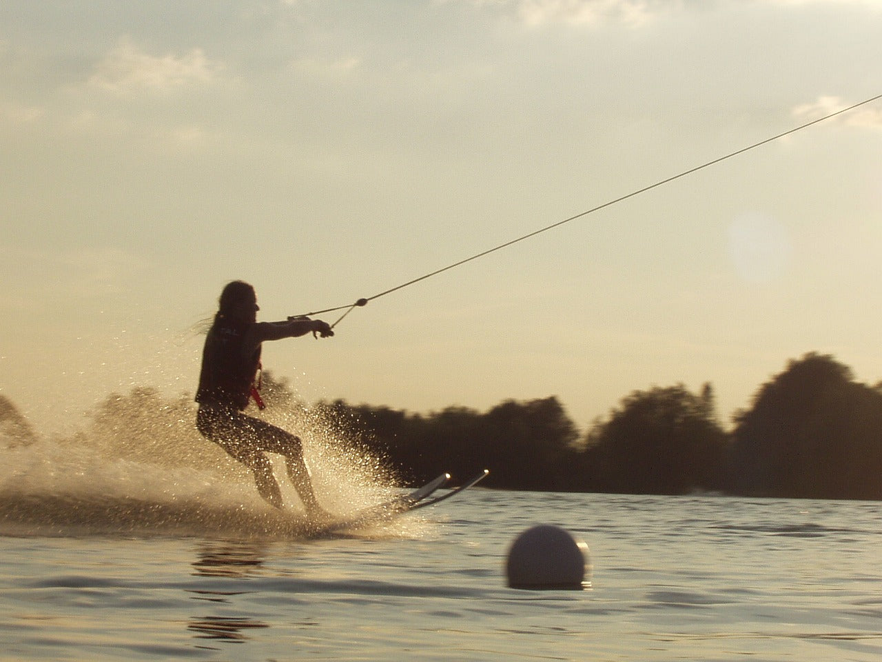 Eine Person fährt Wasserski bei Sonnenuntergang, wird von einem Seil gezogen und erzeugt eine Gischt auf der Wasseroberfläche. Im Vordergrund treibt eine weiße Boje, im Hintergrund sind Bäume silhouettenhaft zu erkennen.