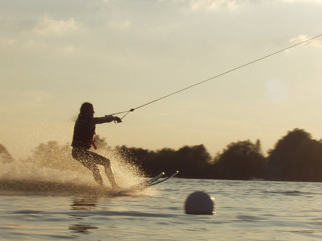 Eine Person fährt Wasserski bei Sonnenuntergang, wird von einem Seil gezogen und erzeugt eine Gischt auf der Wasseroberfläche. Im Vordergrund treibt eine weiße Boje, im Hintergrund sind Bäume silhouettenhaft zu erkennen.