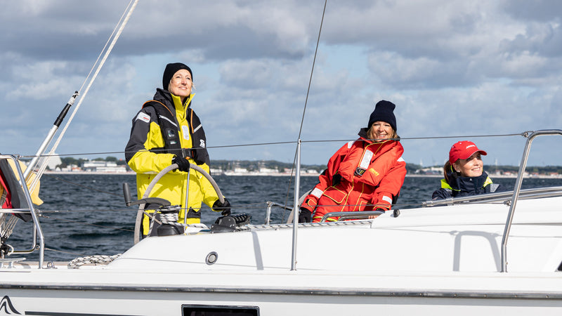Drei Personen in wetterfester Segelkleidung auf einem weißen Segelboot bei bewölktem Himmel und ruhiger See.